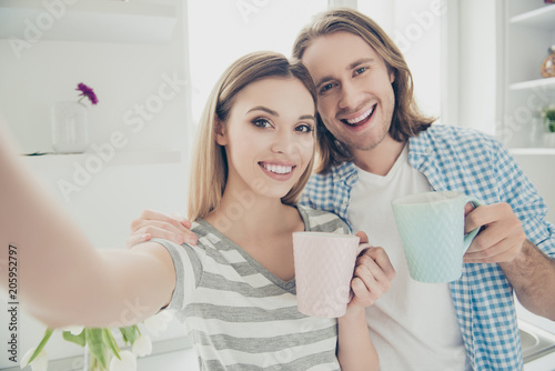 Self portrait of cheerful positive partners drinking hot beverage holding cups in hands shooting selfie on front camera enjoying morning together