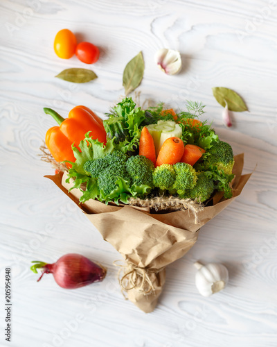 Bouquet of fresh vegetables lies on a white wooden table. Nearby is a red onion  tomatoes  garlic  bay leaf. View from above