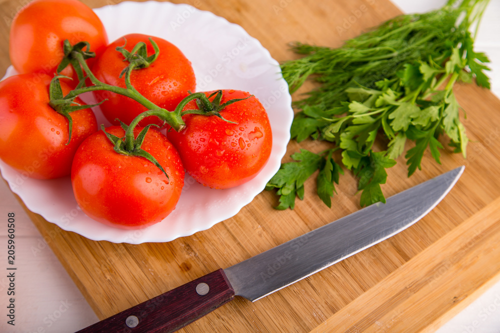 Fresh red tomatoes close up on a white plate at a wooden board with green parsley and a knife Fresh red tomatoes close up on a white plate at a wooden board with green parsley and a knife