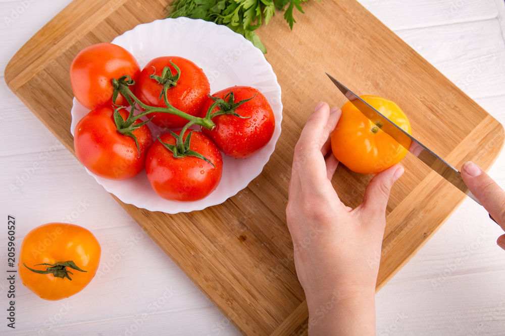 Hands cutting yellow tomato near a white plate with fersh red tomatoes on a wooden board at a white table