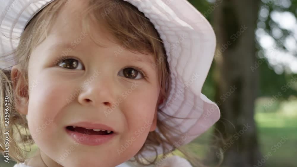 SLOW MOTION, PORTRAIT, CLOSE UP, DOF: Cheerful girl toddler looking around the lovely green tree avenue. Adorable shot of curious little girl wearing a cute white hat observing the tranquil park.