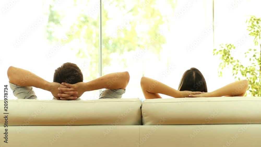 Rear view of a couple relaxing on a sofa at home and looking outside a green background through the window of the living room
