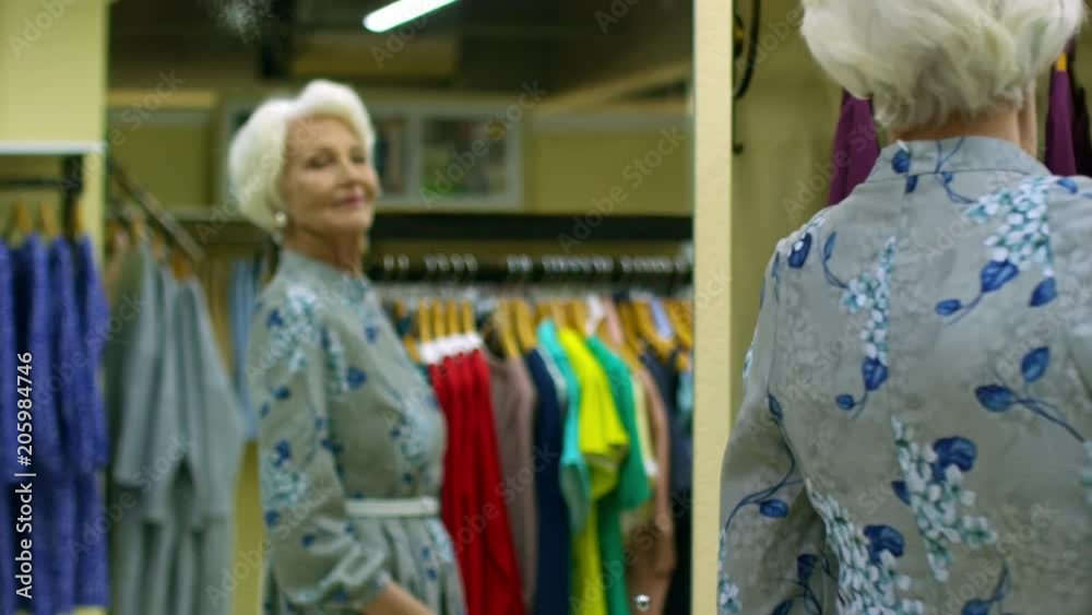 Medium shot of elegant retired woman in pearl earrings posing in front of mirror in new floral dress Medium shot of elegant retired woman in pearl earrings posing in front of mirror in new floral dress