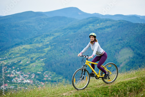 Sporty girl riding on yellow bike on rural trail on high mountain range at cloudy evening, wearing helmet and violet leggins. Forests and small city on blurred background. Outdoor sport activity.