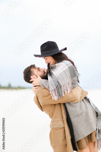 Caucasian man with beard picking up young woman wearing hat, coat and grey scarf on white snow background. Concept of gladden couple and seasonal photo session. photo