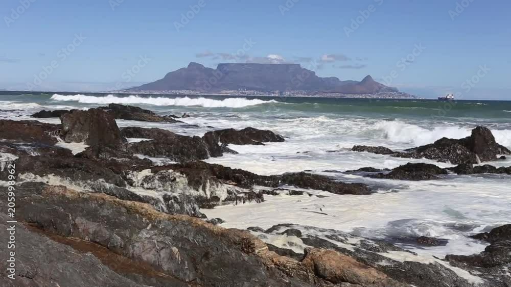 Daytime view of table mountain across table bay as waves crash ashore on the rocks in Cape Town South Africa with large rock in foreground Daytime view of table mountain across table bay as waves crash ashore on the rocks in Cape Town South Africa with large rock in foreground