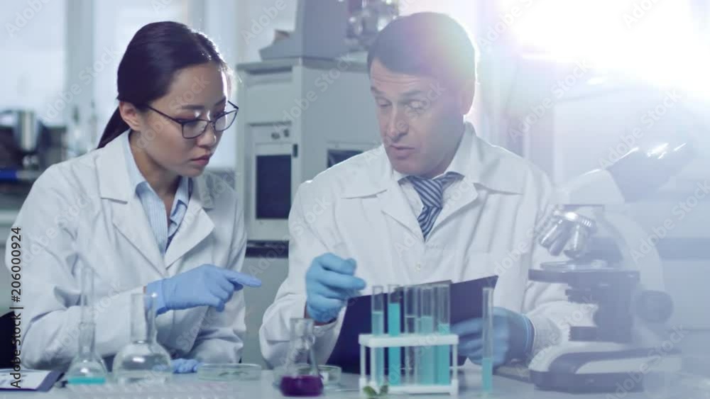 Tracking shot of two scientists studying liquids in test tubes and comparing test results with data they have on clipboard Tracking shot of two scientists studying liquids in test tubes and comparing test results with data they have on clipboard