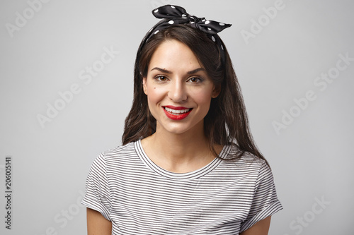 Beauty, fashion and cosmetics concept. Isolated shot of beautiful gorgeous young female with charming smile posing in studio, wearing bright make up, red lipstick, headband and sailor shirt