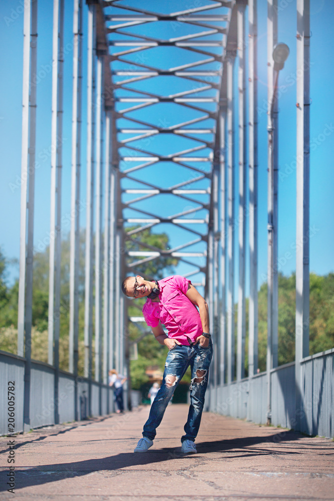 Cute emotional man with headphones in glasses in a pink shirt walking on a pedestrian bridge Cute emotional man with headphones in glasses in a pink shirt walking on a pedestrian bridge