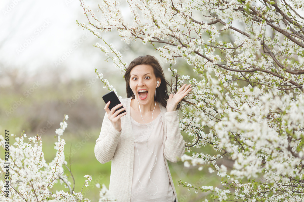 Young overjoyed beautiful woman in casual clothes with earphones listening music holding mobile phone spreading hands in garden or park on blooming tree background. Spring flowers. Lifestyle concept. Young overjoyed beautiful woman in casual clothes with earphones listening music holding mobile phone spreading hands in garden or park on blooming tree background. Spring flowers. Lifestyle concept.