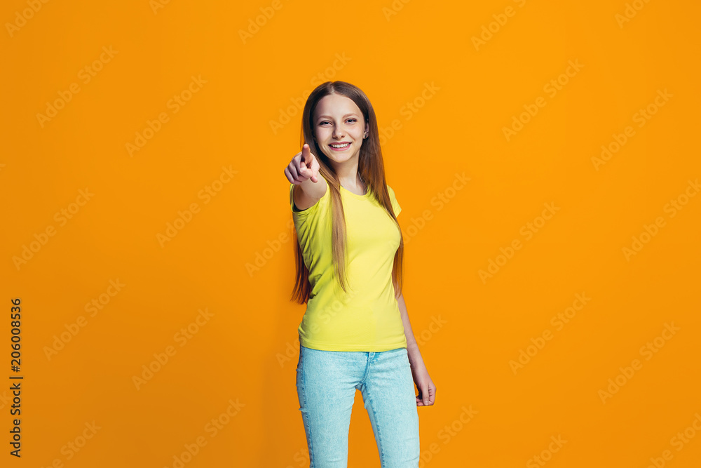 The happy teen girl pointing to you, half length closeup portrait on orange background. The happy teen girl pointing to you, half length closeup portrait on orange background.