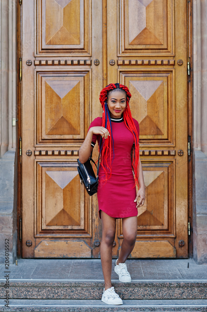 Cute and slim african american girl in red dress with dreadlocks and backpack posed against school large wooden door. Stylish black student. Stock Foto Adobe Stock