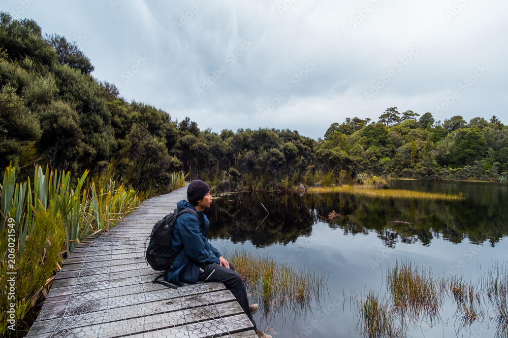 A young Asian man sitting on the path beside a beautiful lake in a cloudy day.