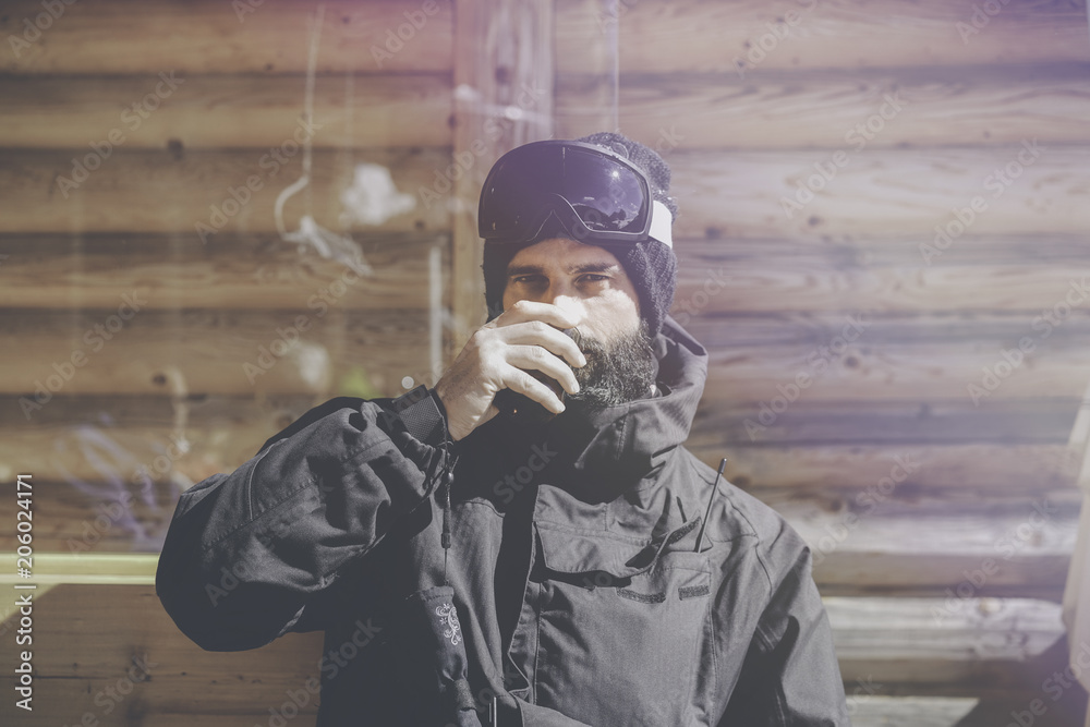Bearded handsome snowboarded taking rest after ride session.Young man drinking cup of hot tea on sunny terrace. Blurred background.Horizontal.Visual effects Bearded handsome snowboarded taking rest after ride session.Young man drinking cup of hot tea on sunny terrace. Blurred background.Horizontal.Visual effects