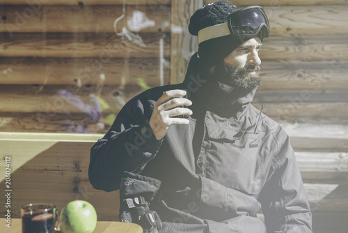 Bearded handsome snowboarded taking rest after ride session.Young man drinking cup of hot tea on sunny terrace. Blurred background.Horizontal.Visual effects photo