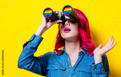 Young pink hair girl in blue shirt holding a binoculars with rainbow. Portrait on isolated yellow background