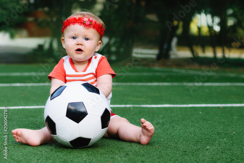 Little girl in sport shirt and red hair band playing with a soccer ball at football field. Summer kid sport concept