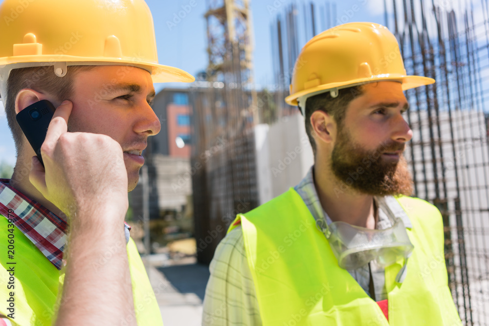Side view close-up of a worker wearing yellow hard hat, while talking on mobile phone during work on the construction site of a building in progress Side view close-up of a worker wearing yellow hard hat, while talking on mobile phone during work on the construction site of a building in progress