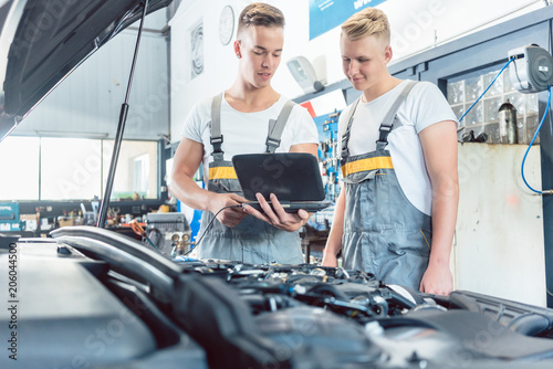 Experienced auto mechanic using a laptop for scanning and interpreting engine error codes next to an apprentice in a modern automobile repair shop photo
