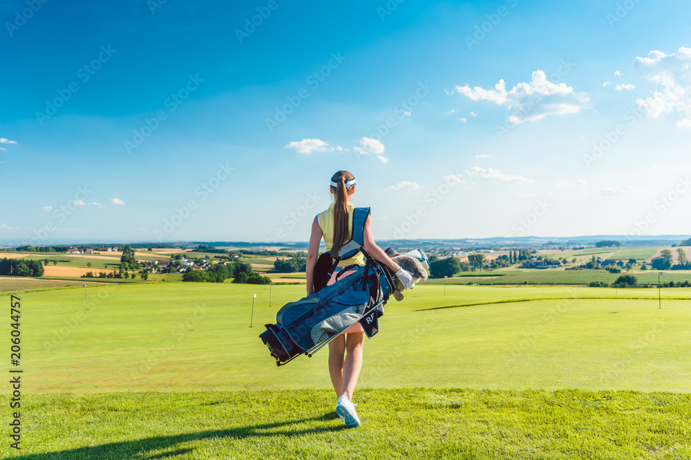 Full length rear view of an active woman carrying a blue stand bag, while walking on the grass towards a training area for professional golf in a sunny day Full length rear view of an active woman carrying a blue stand bag, while walking on the grass towards a training area for professional golf in a sunny day