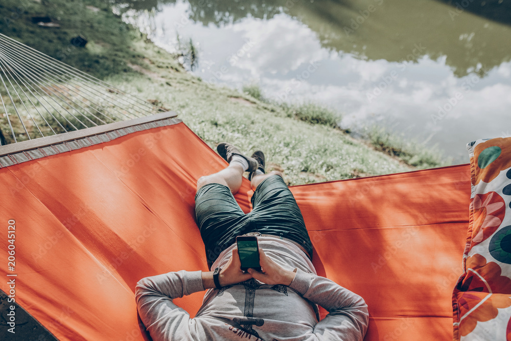 hipster man holding smart phone with empty screen with space for text, and relaxing in hammock in sunny summer park. mock up. guy looking at blank phone, technology communication concept