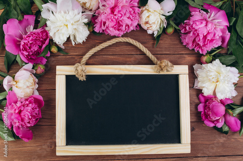Pink and white peonies around blackboard on a wooden background. Copy space and flat lay.