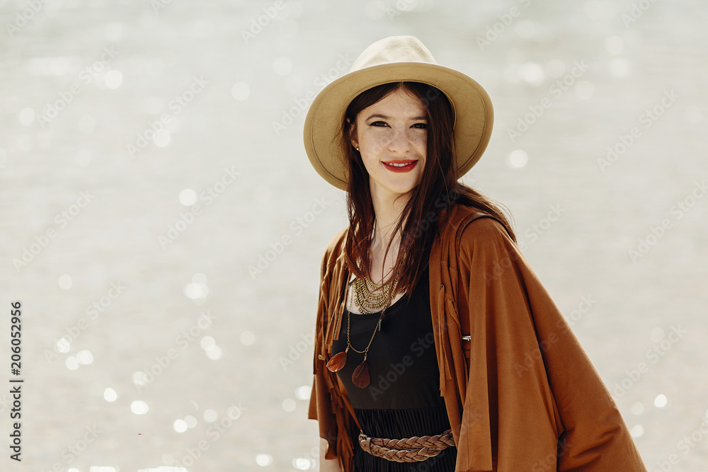 stylish boho traveler woman in hat, fringe poncho posing near water river beach, gypsy hipster girl look, summer travel. atmospheric moment. space for text. wanderlust
