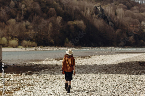 stylish boho traveler woman in hat back view, fringe poncho posing near water river beach in mountains, gypsy hipster girl. wanderlust summer travel. atmospheric moment. space for text.
