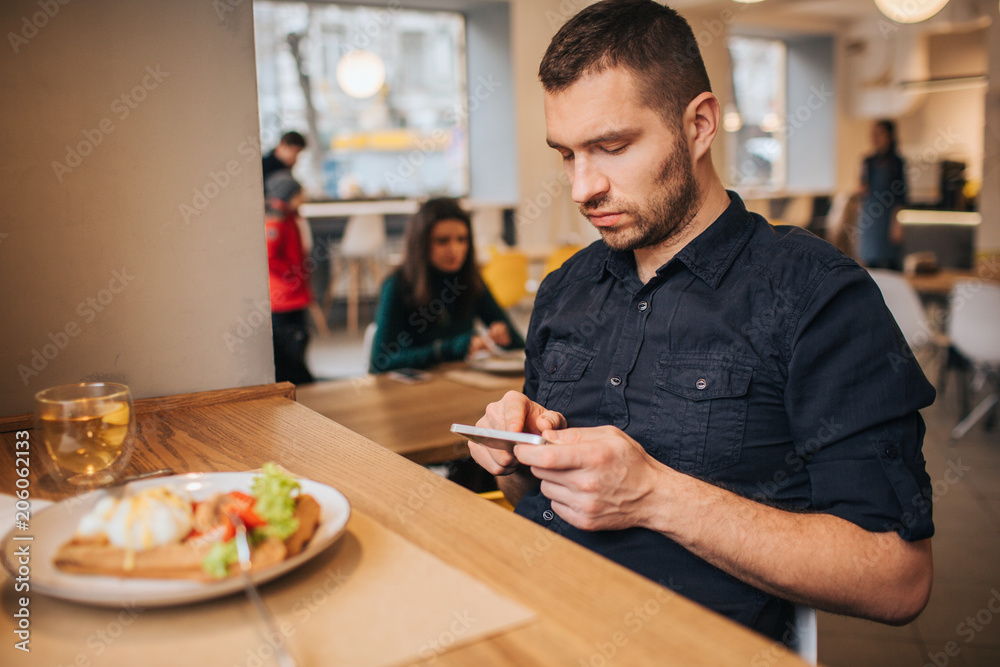 Calm and serious guy is sitting at table and playing with his phone. He is concentrated on that. There are plate with food and cup of tea at the table. Calm and serious guy is sitting at table and playing with his phone. He is concentrated on that. There are plate with food and cup of tea at the table.