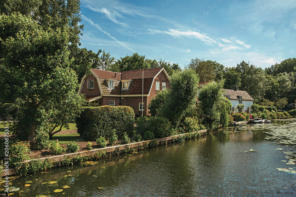 Fototapeta premium Tree-lined canal with aquatic plants, boats on the banks and brick houses on a sunny day in Weesp. Quiet and pleasant village full of canals and green near Amsterdam. Northern Netherlands.