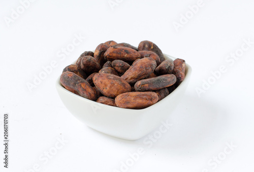 Pile of raw organic unpeeled cacao beans in a small bowl, isolated on white background.