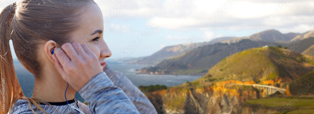 fitness, sport and technology concept - close up of happy woman listening to music in earphones over bixby creek bridge on big sur coast of california background