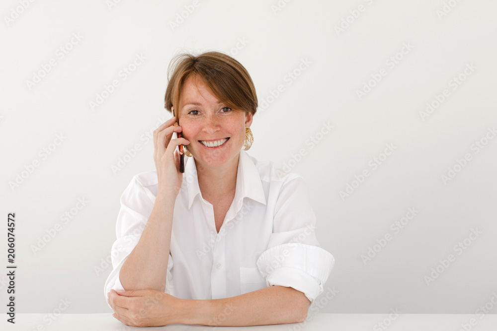 Beautiful adult woman in white shirt speaking on smartphone sitting at desk on white and smiling at camera. Beautiful adult woman in white shirt speaking on smartphone sitting at desk on white and smiling at camera.