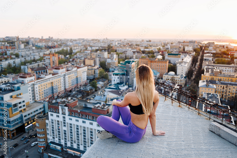 Young girl in sports uniform sits on the edge of the roof during sunset and looks to the horizon.