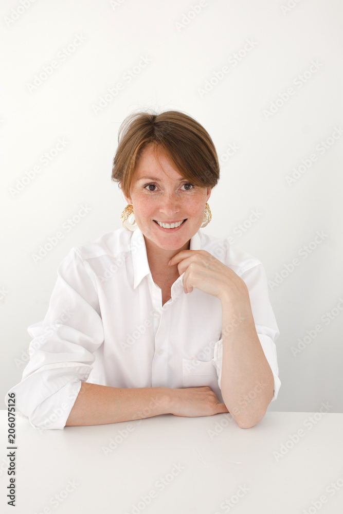 Beautiful adult woman in white shirt sitting at desk on white and smiling at camera Beautiful adult woman in white shirt sitting at desk on white and smiling at camera
