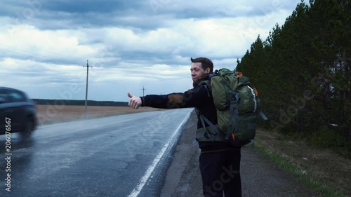 A tourist hitchhiker walks along the road with a backpack on his shoulders, lifting his thumb up. Traveler is trying to stop the car. The weather is cloudy and rainy.