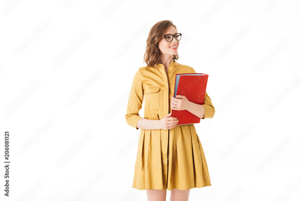 Portrait of nice smiling lady in eyeglasses and yellow dress standing with red folder in hand and looking aside on white background isolated