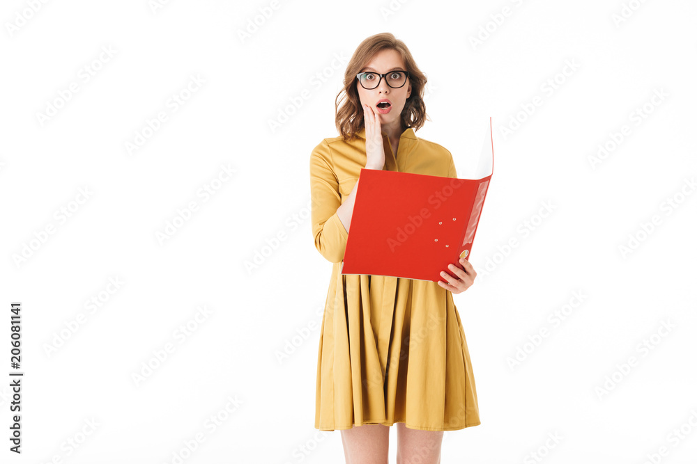 Portrait of young lady in eyeglasses and yellow dress holding red folder while amazedly looking in camera on white background isolated Portrait of young lady in eyeglasses and yellow dress holding red folder while amazedly looking in camera on white background isolated