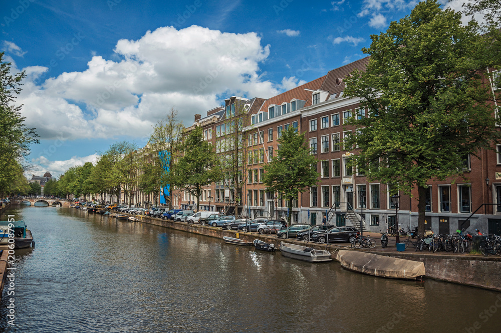 Tree-lined canal with old brick buildings, steeple, moored boats and sunny blue sky in Amsterdam. The city is famous for its huge cultural activity, graceful canals and bridges. Northern Netherlands.