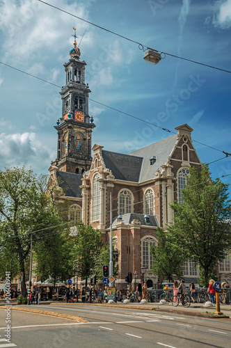 Street with old brick church steeple, trees and people passing by in Amsterdam. Famous for its huge cultural activity, graceful canals and bridges. Northern Netherlands.