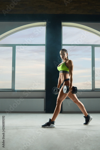 Athletic woman keeping dumbbells in big, spacy hall. Having strong, fit body with heatlthy tanned skin and muscles. Doing fitness exercises. Wearing modern sportswear. Looking at camera.