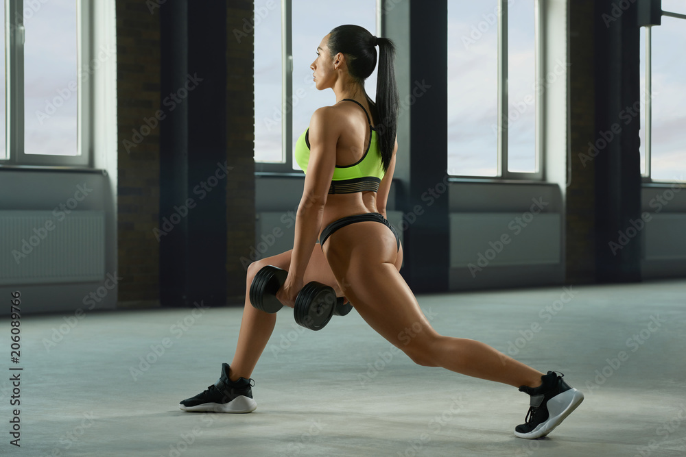 Athletic girl performing fallouts keeping dumbbells in spacy gym with panoramic windows. Having strong, fit body with heatlthy tanned skin, muscles. Doing fitness exercises. Wearing modern sportswear.