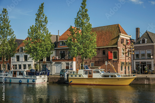 Wide canal with brick houses, boats moored on its bank reflected in water and blue sky of sunset in Weesp. Quiet and pleasant village full of canals and green near Amsterdam. Northern Netherlands.
