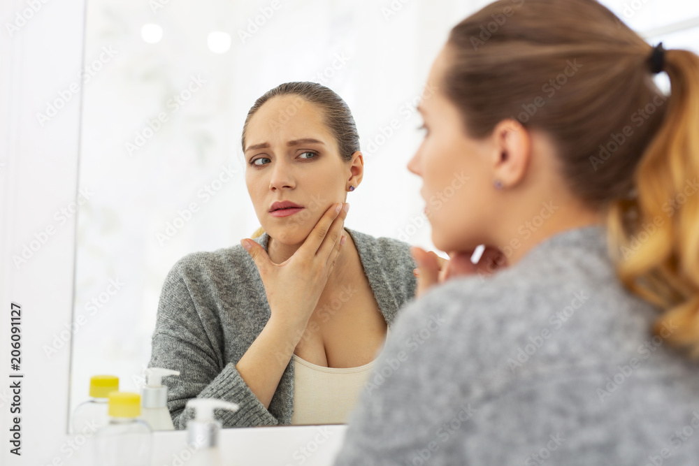 Common problem. Uneasy worried woman looking at mirror and putting hand on neck Common problem. Uneasy worried woman looking at mirror and putting hand on neck