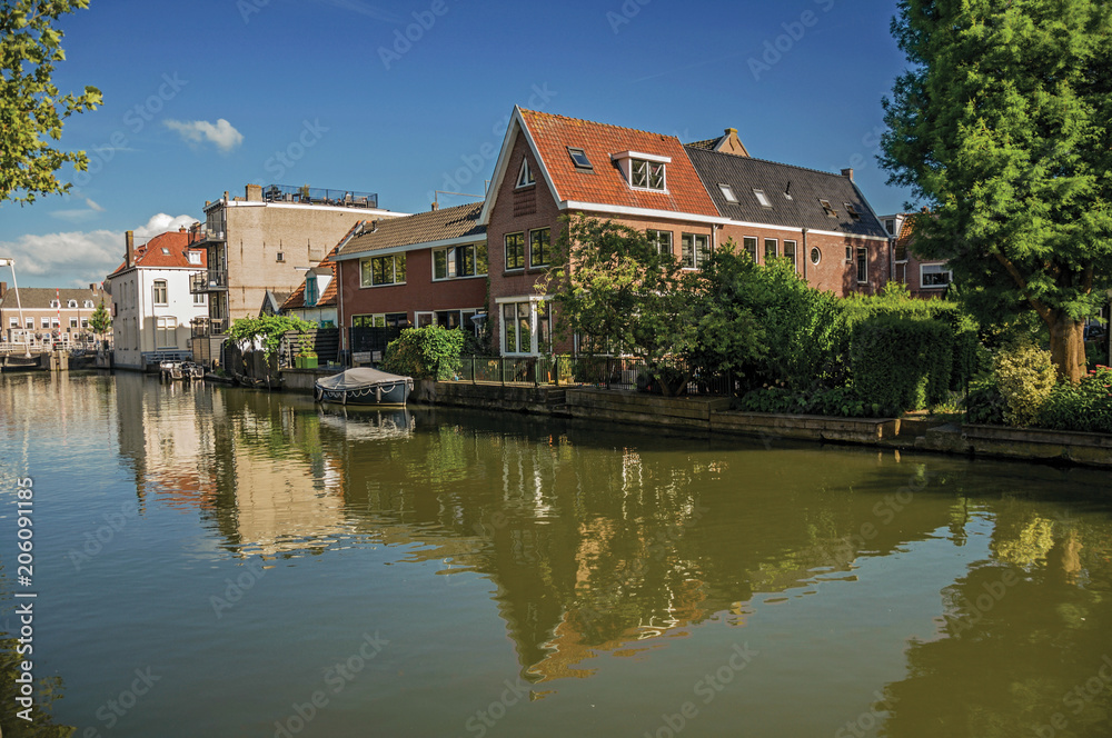 Wide canal with brick houses and boat moored on its bank reflected in water under blue sky of sunset in Weesp. Quiet and pleasant village full of canals and green near Amsterdam. Northern Netherlands.