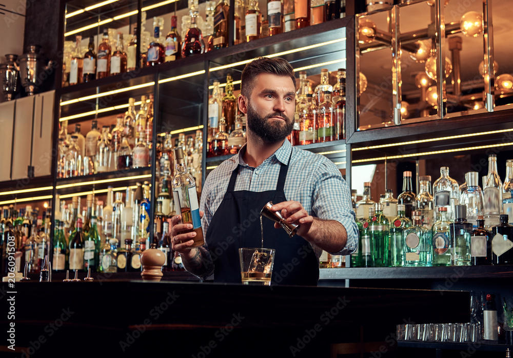 Stylish brutal barman in a shirt and apron makes a cocktail at bar counter background. Stylish brutal barman in a shirt and apron makes a cocktail at bar counter background.