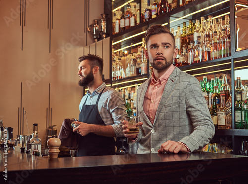 Stylish brutal barman in a shirt and apron makes a cocktail at bar counter background.