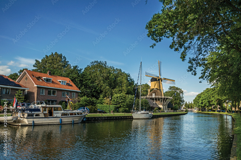 Boats and wooden yellow windmill next to wide tree-lined canal on the bright light of sunset at Weesp. Quiet and pleasant village full of canals and green near Amsterdam. Northern Netherlands. Boats and wooden yellow windmill next to wide tree-lined canal on the bright light of sunset at Weesp. Quiet and pleasant village full of canals and green near Amsterdam. Northern Netherlands.