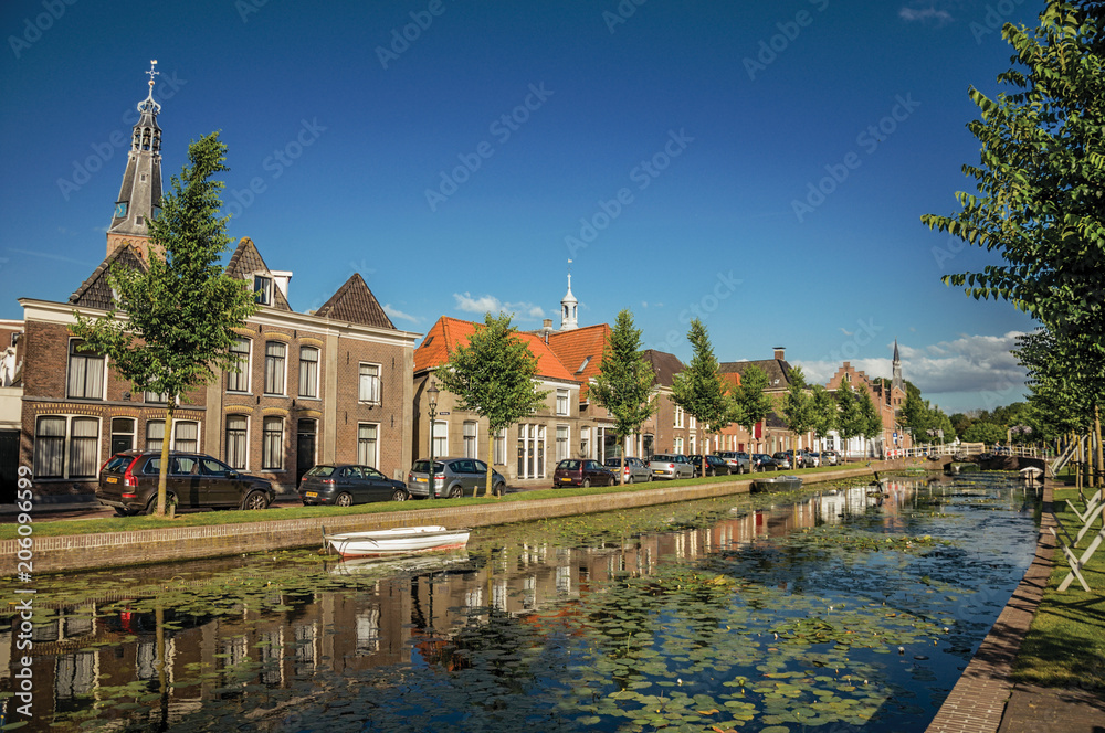 Tree-lined canal with boat, church bell tower and brick houses at the bank on sunset in Weesp. Quiet and pleasant village full of canals and green near Amsterdam. Northern Netherlands.