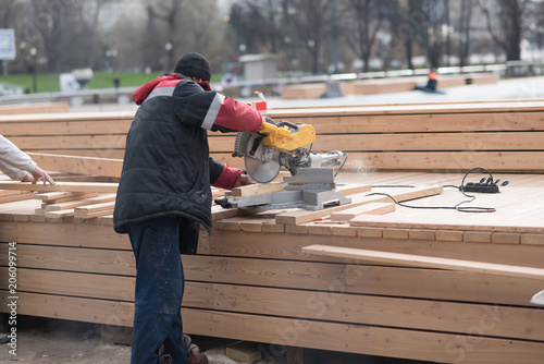 Man in a sweatshirt and pants cutting huge piece of wood by electric circular saw. outdoor